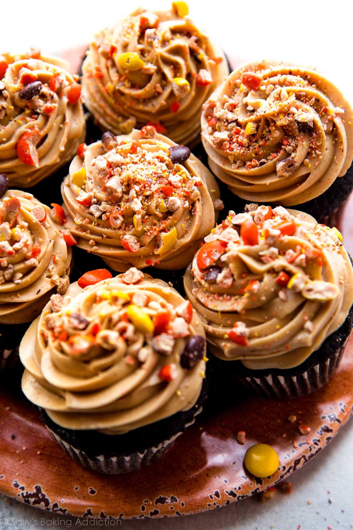 dark chocolate cupcakes on an orange plate topped with swirls of peanut butter frosting and crushed Reese's Pieces candies