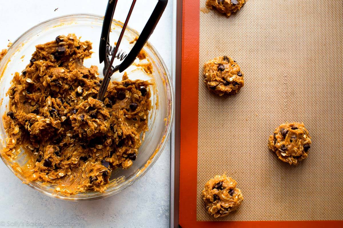 pumpkin oatmeal chocolate chip cookie dough in a glass bowl with a cookie scoop and a baking sheet with cookie dough before baking