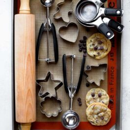 overhead image of a wood rolling pin, cookie scoops, measuring cups, cookie cutters, and cookies on a baking sheet lined with a silpat baking mat