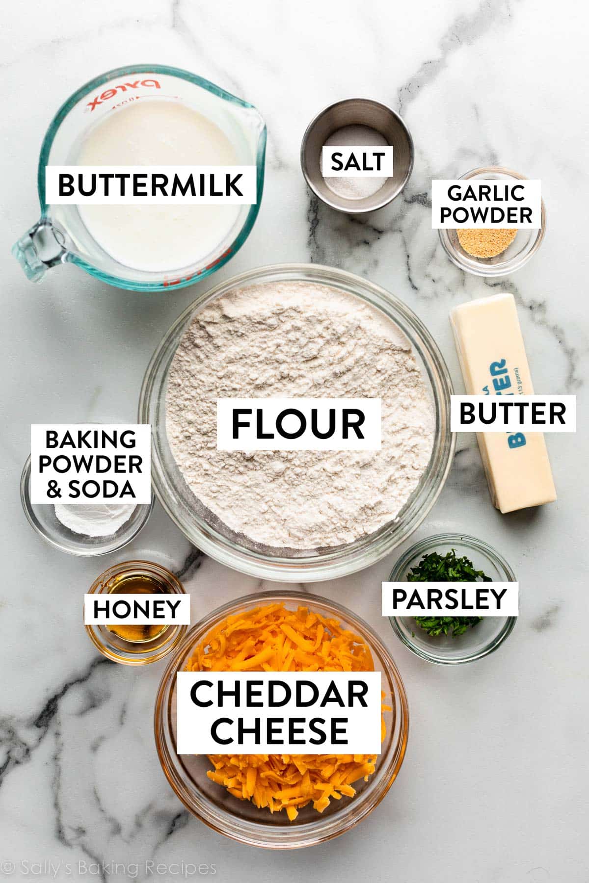 ingredients on marble counter including bowl of flour, cheddar cheese, parsley, garlic powder, salt, a stick of butter, and a measuring cup of milk.