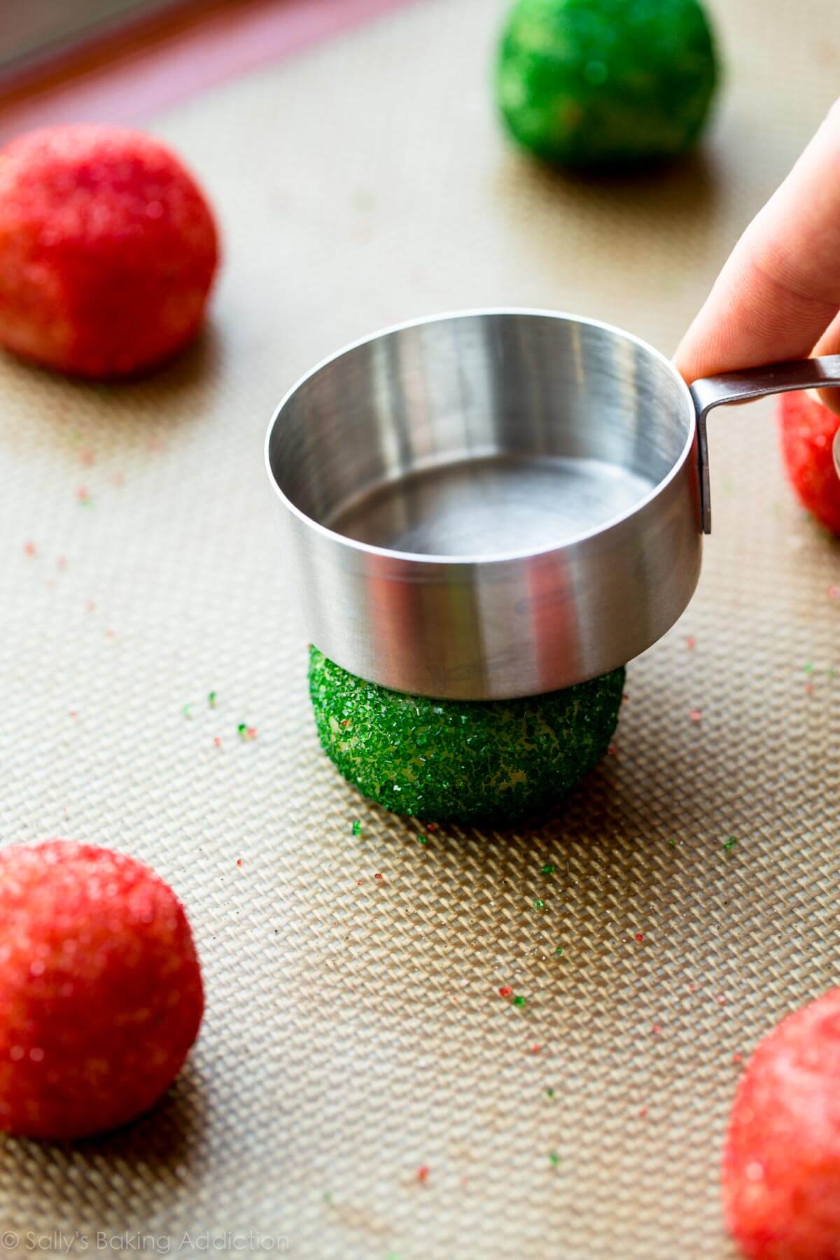 Christmas sugar cookie dough rolled in green and red sprinkles and a measuring cup flattening down the top of a dough ball