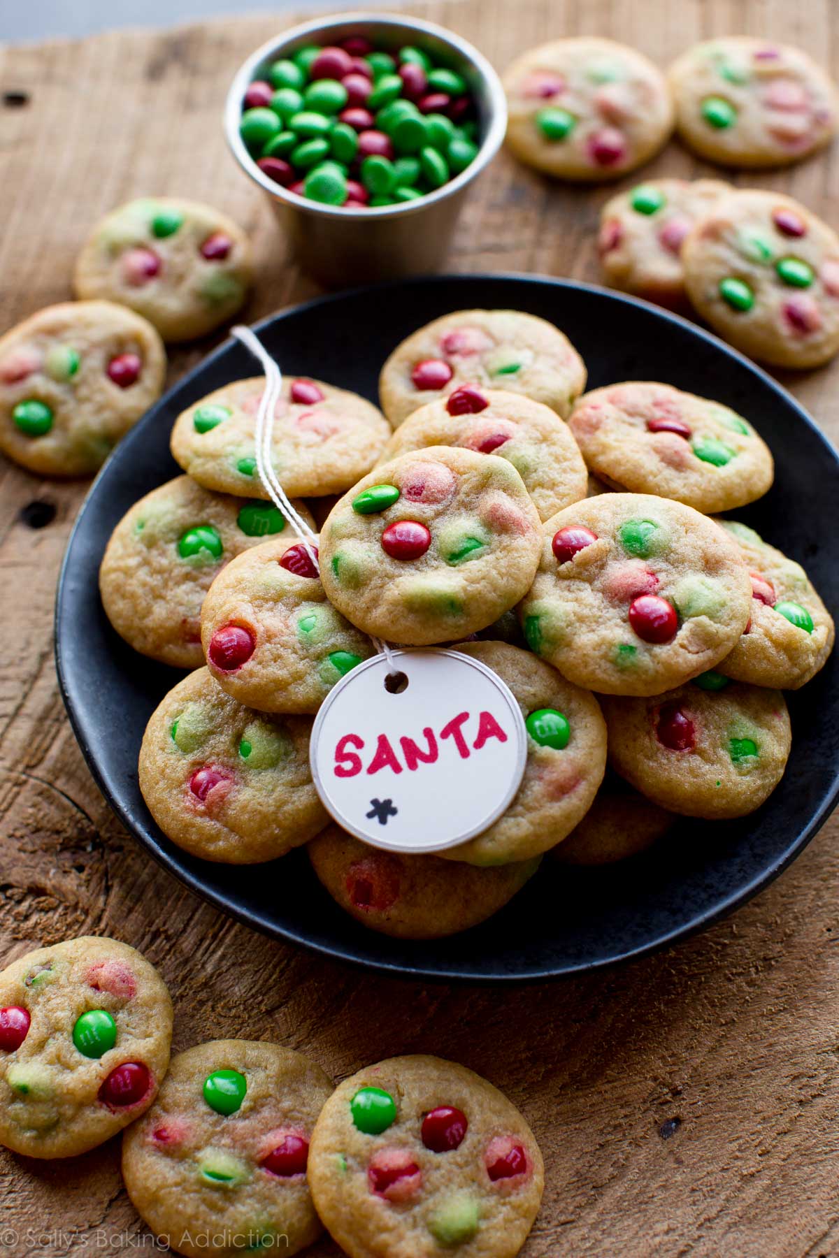 mini M&M cookies with Christmas M&Ms on a black plate with a Santa tag