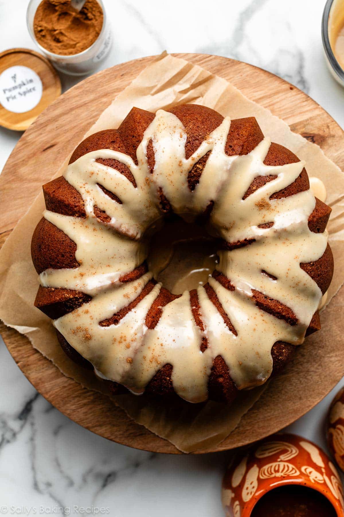 overhead shot of pumpkin Bundt cake on wooden cake stand with maple icing on top and a sprinkle of pumpkin pie spice.