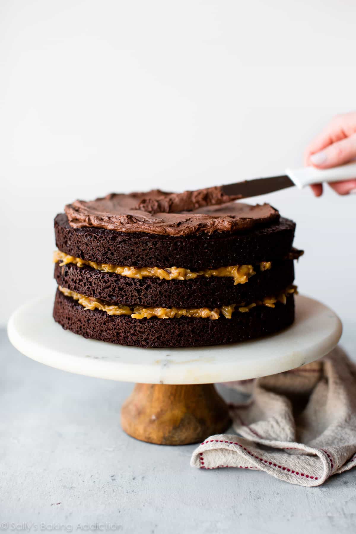 spreading frosting onto German chocolate cake on a marble and wood cake stand