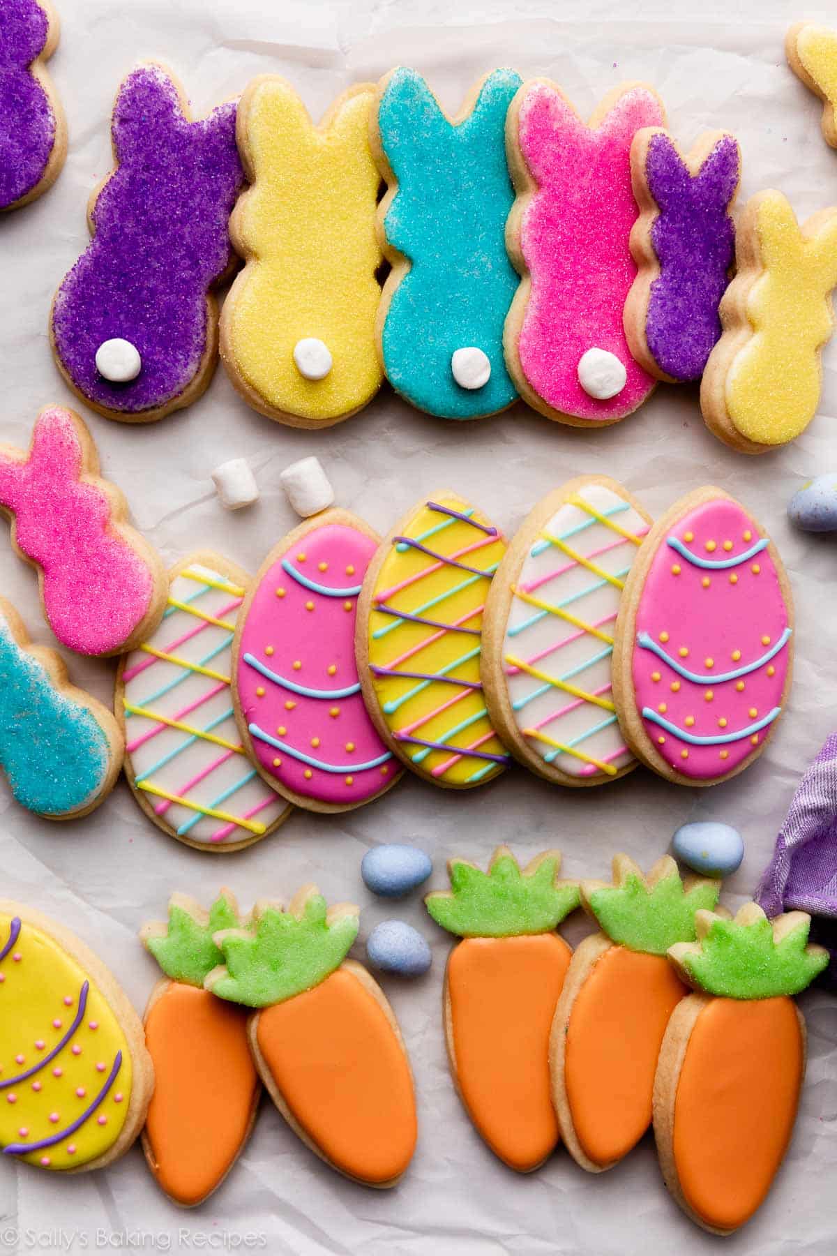 decorated Easter cookies on parchment paper including bunnies with marshmallow tails, Easter eggs, and carrots with orange and green icing.