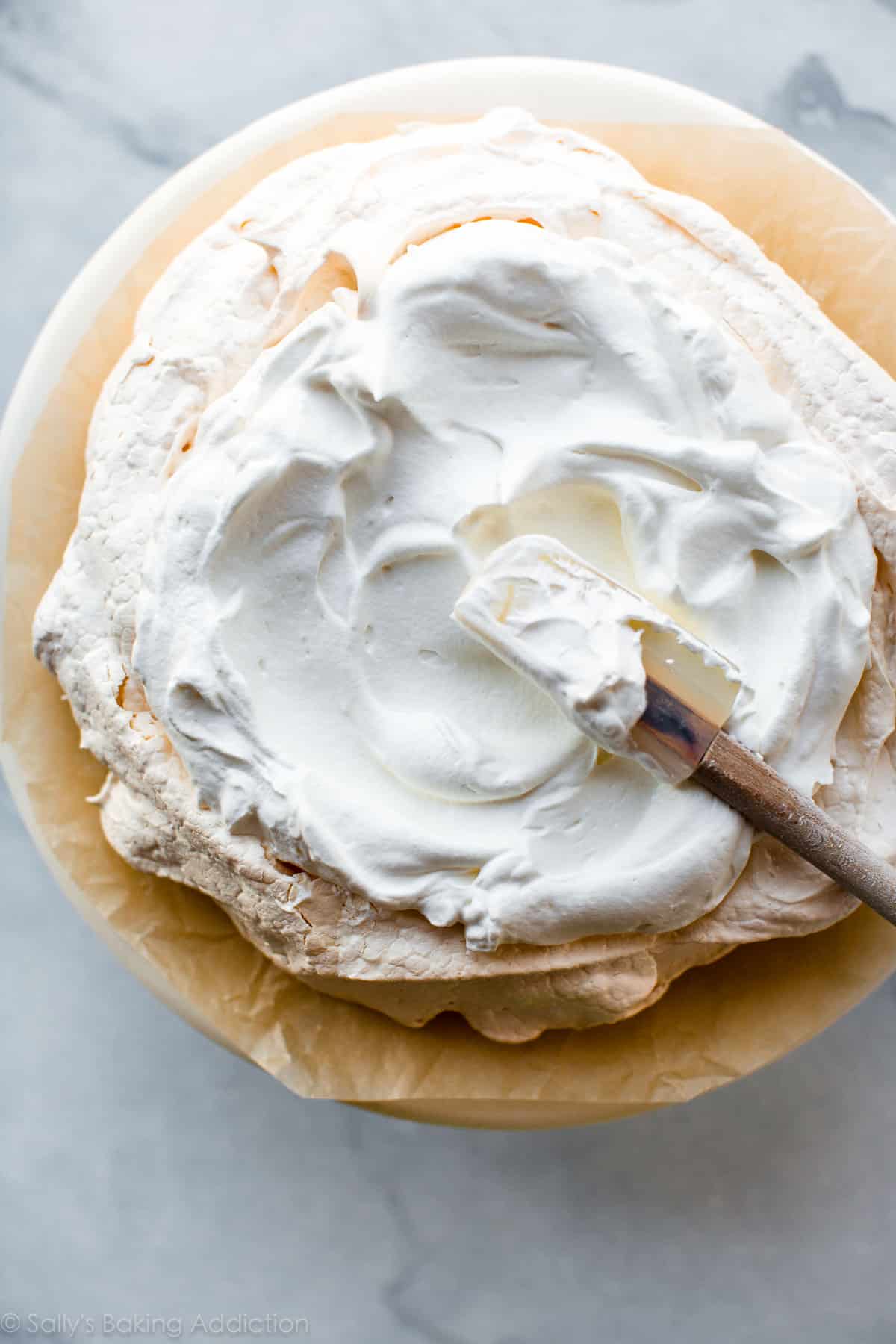 overhead image of spreading whipped cream onto baked pavlova on a marble cake stand