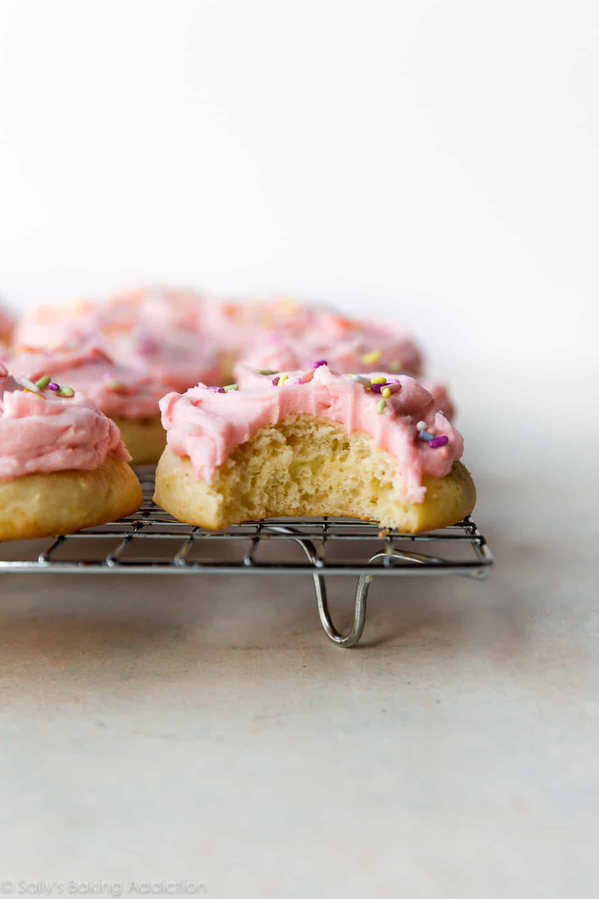 side view of a sugar cookie with a bite taken from it topped with pink buttercream and sprinkles on a cooling rack