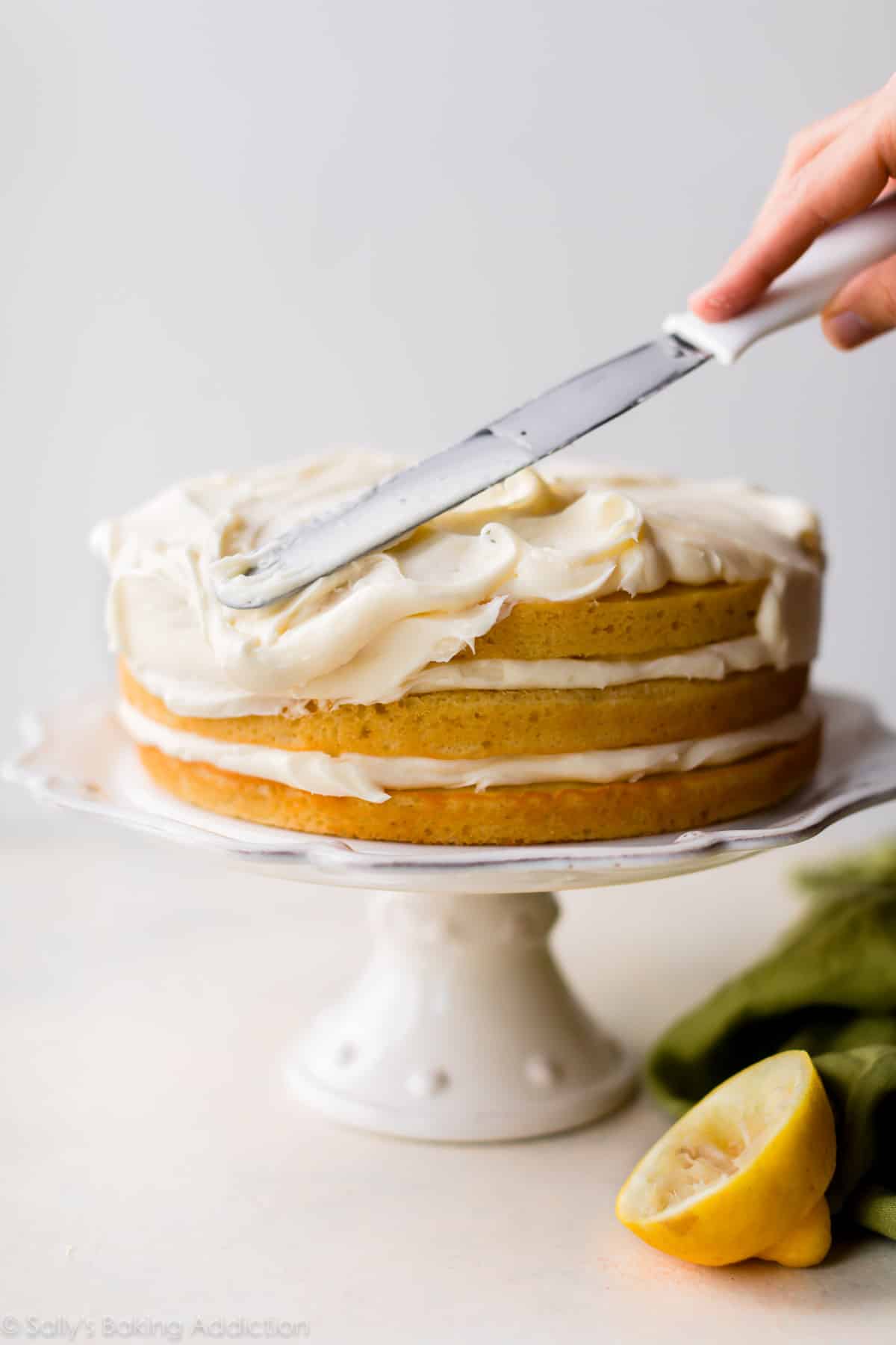 spreading frosting onto lemon cake on a white cake stand