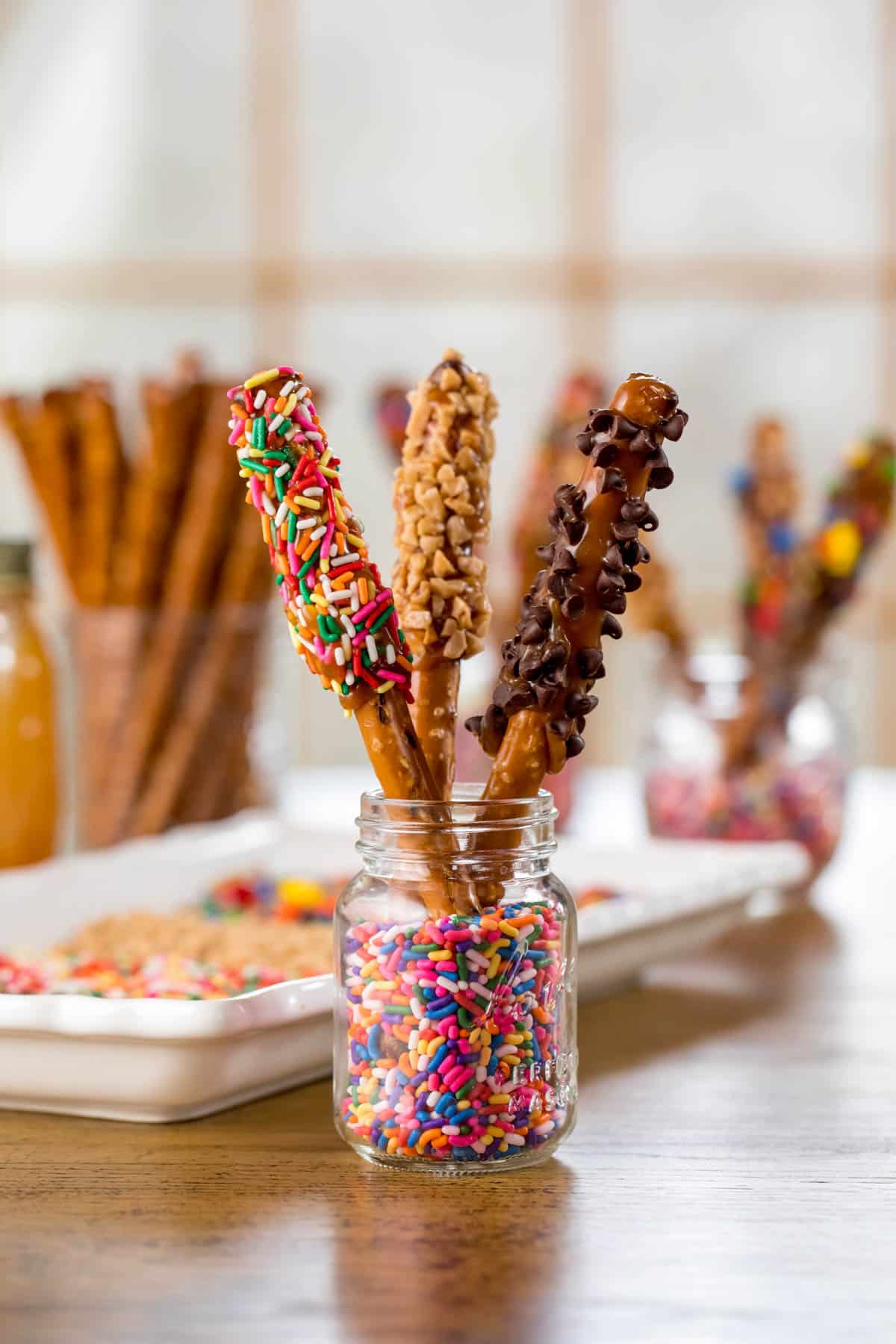 3 pretzel rods dipped in caramel and various toppings standing up in a glass jar with sprinkles