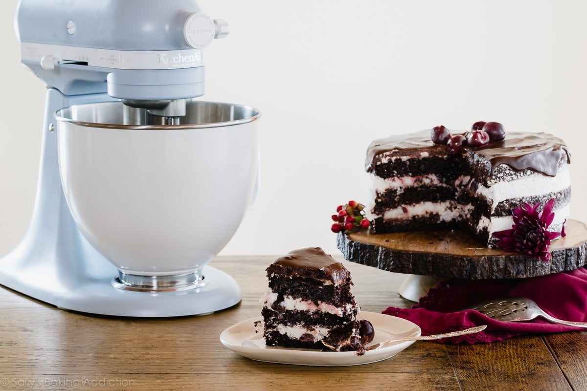 slice of black forest cake on a white plate, the rest of the cake on a wood slice cake stand, and a blue stand mixer