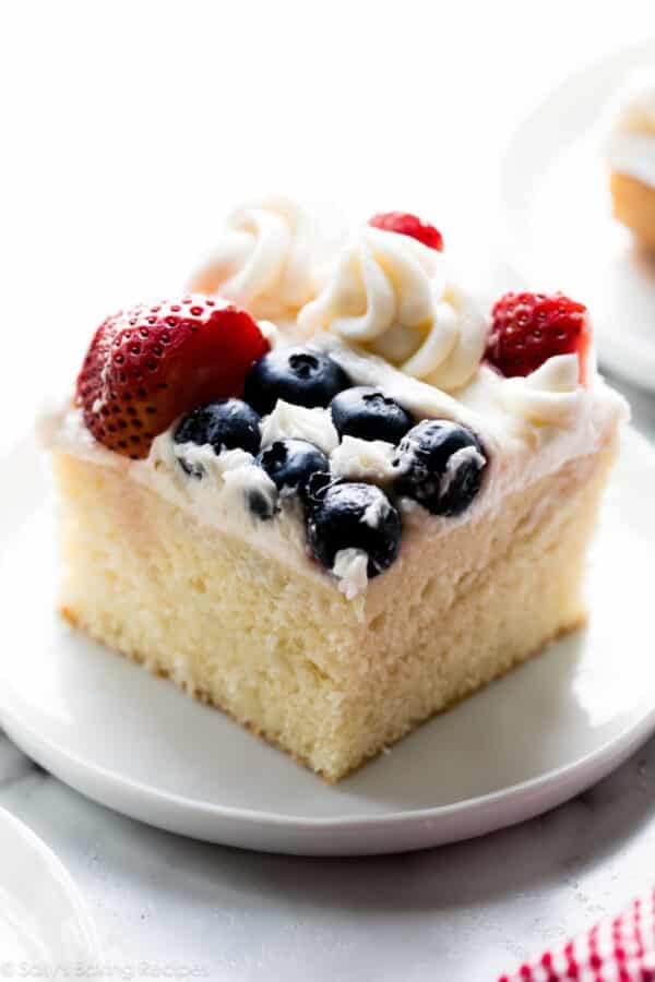 square slice of American flag cake on white plate with blueberries and strawberries on top.