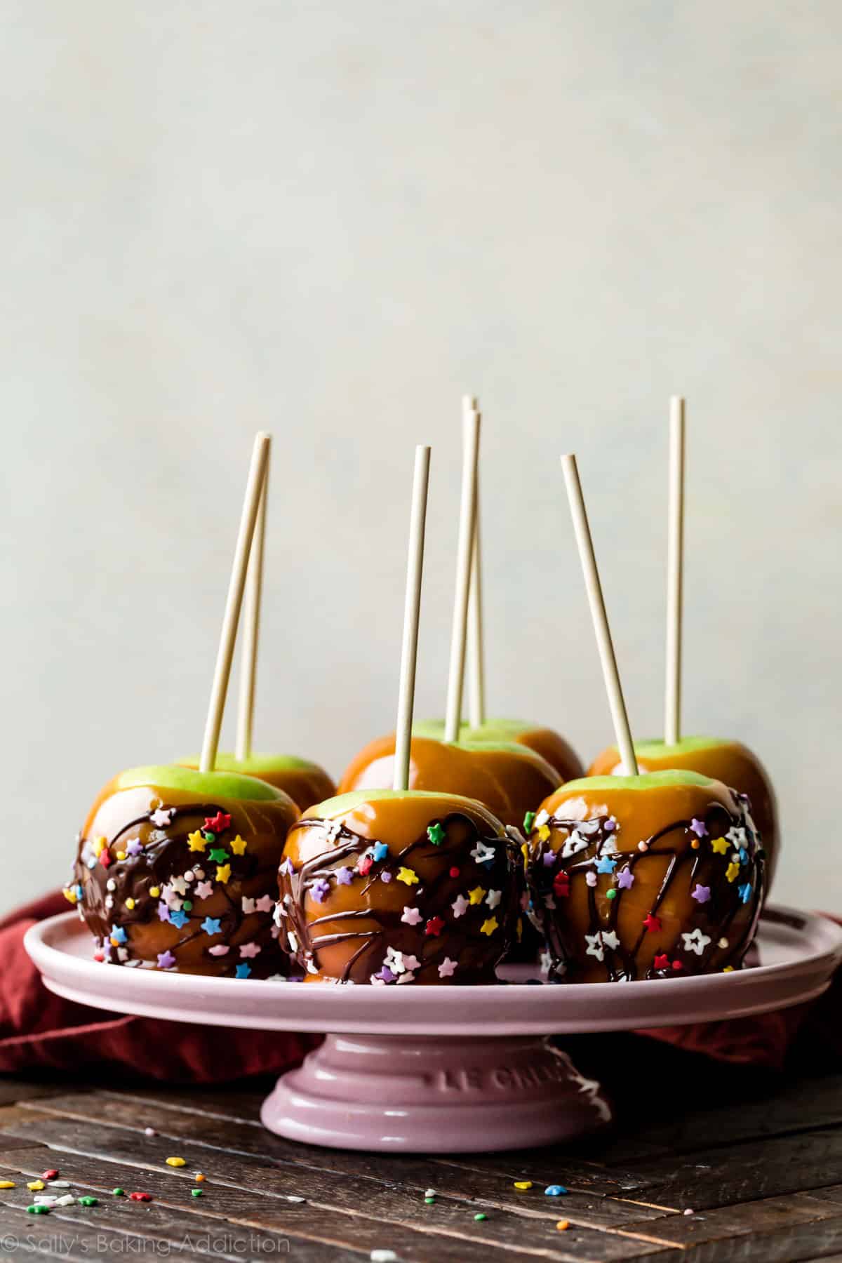 caramel apples with sprinkles and chocolate drizzle on pink cake stand