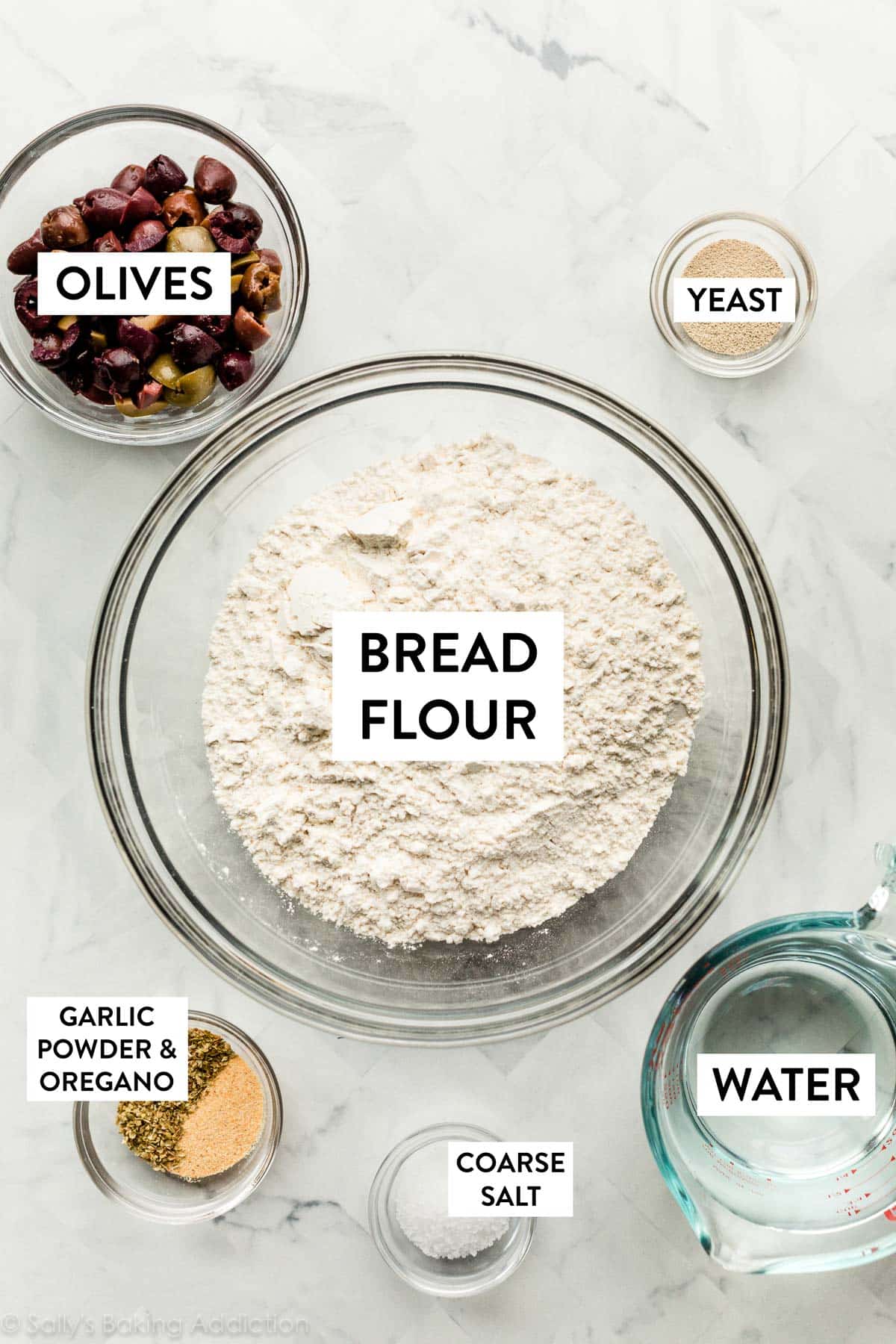ingredients in bowls including bread flour, olives, yeast, water, coarse salt, garlic powder, and dried oregano