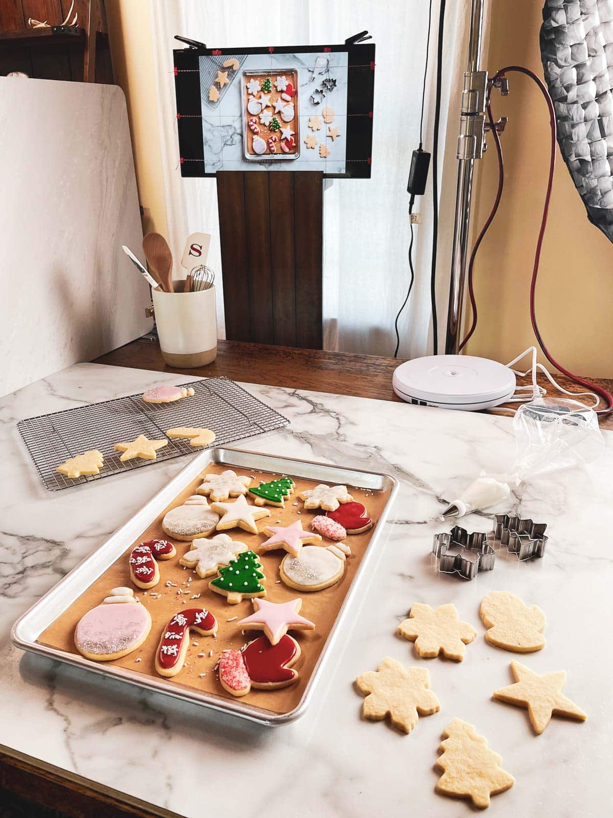 decorated Christmas sugar cookies on marble board with cookie cutters and monitor screen behind it.
