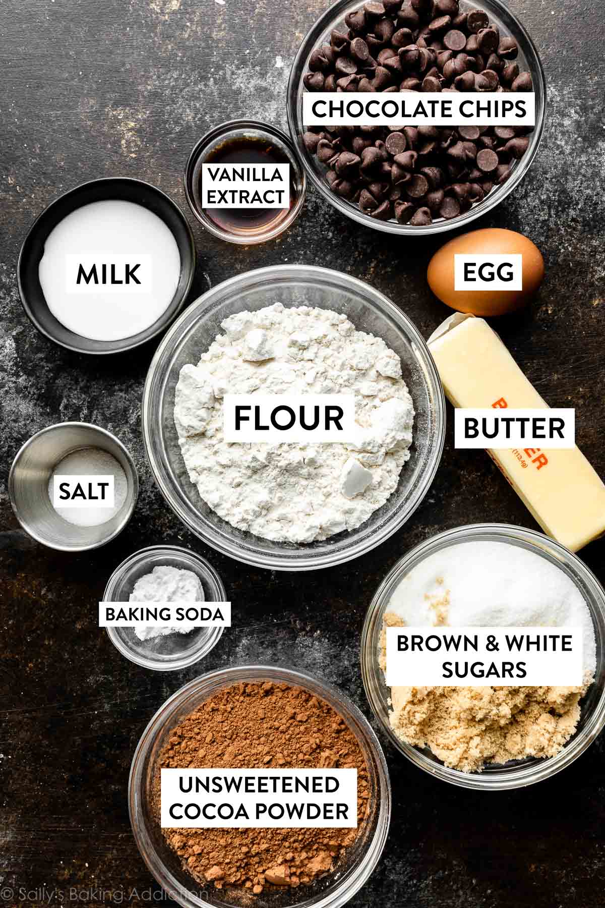 farina, burro, uova, gocce di cioccolato, latte, vaniglia, sale e altri ingredienti sul bancone nero.