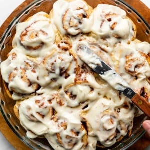 spreading cream cheese icing on a round pan of cinnamon rolls.