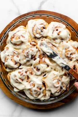 spreading cream cheese icing on a round pan of cinnamon rolls.