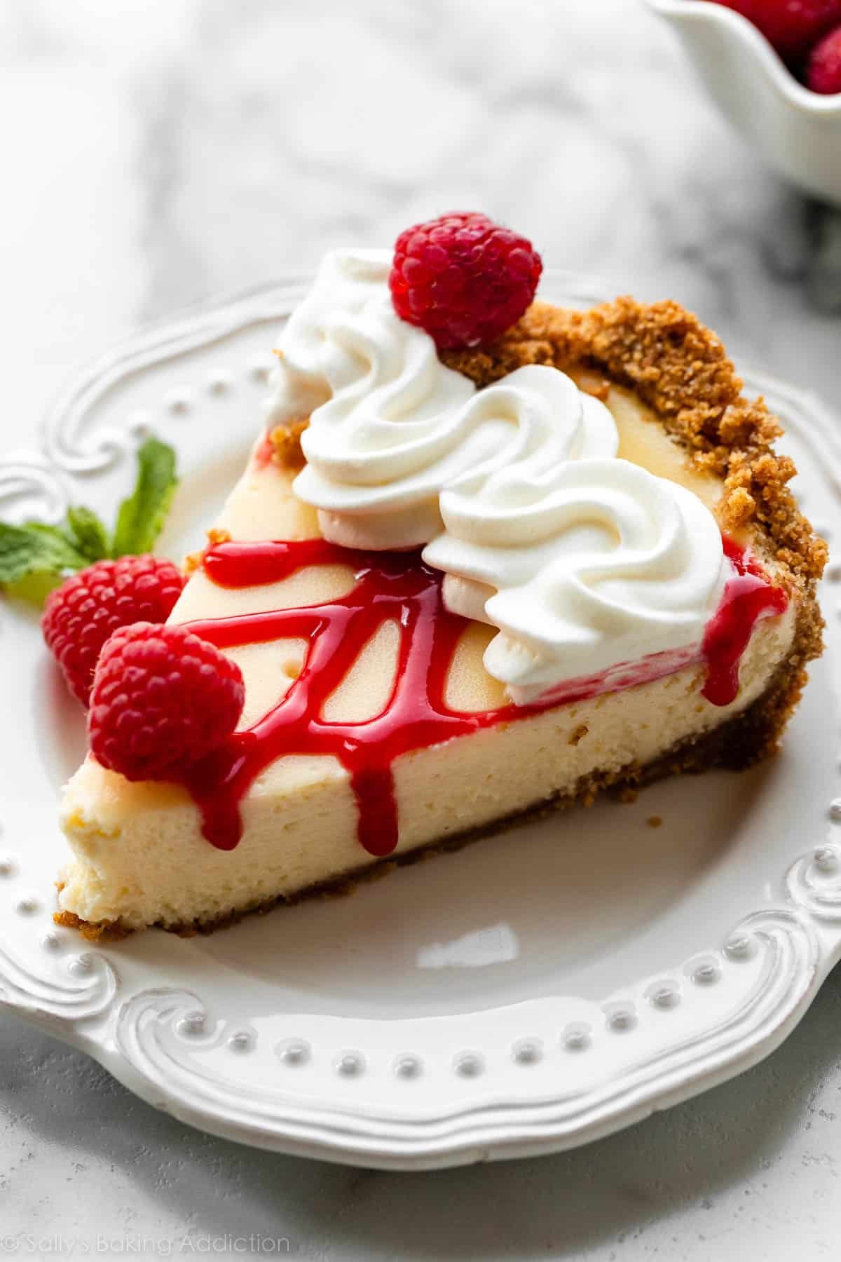 slice of cheesecake pie on white plate with whipped cream, raspberry sauce, and fresh raspberries on top.