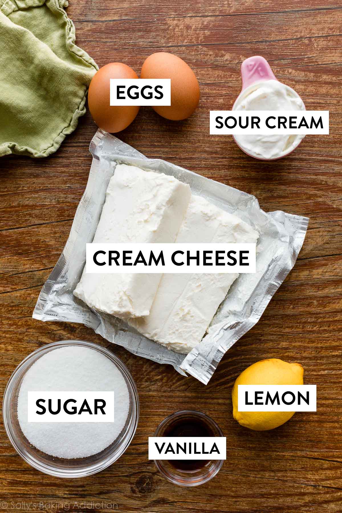 cream cheese, lemon, vanilla extract, sugar, eggs, and sour cream on brown wood countertop.