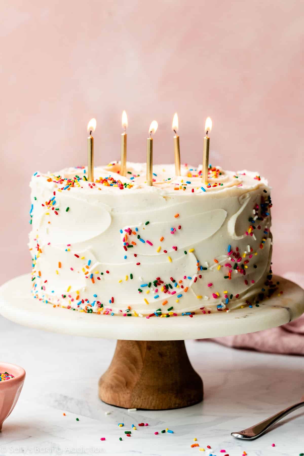 Confetti birthday layer cake with sprinkles and 5 gold, lit candles on top sitting on a marble cake stand with a pink backdrop.