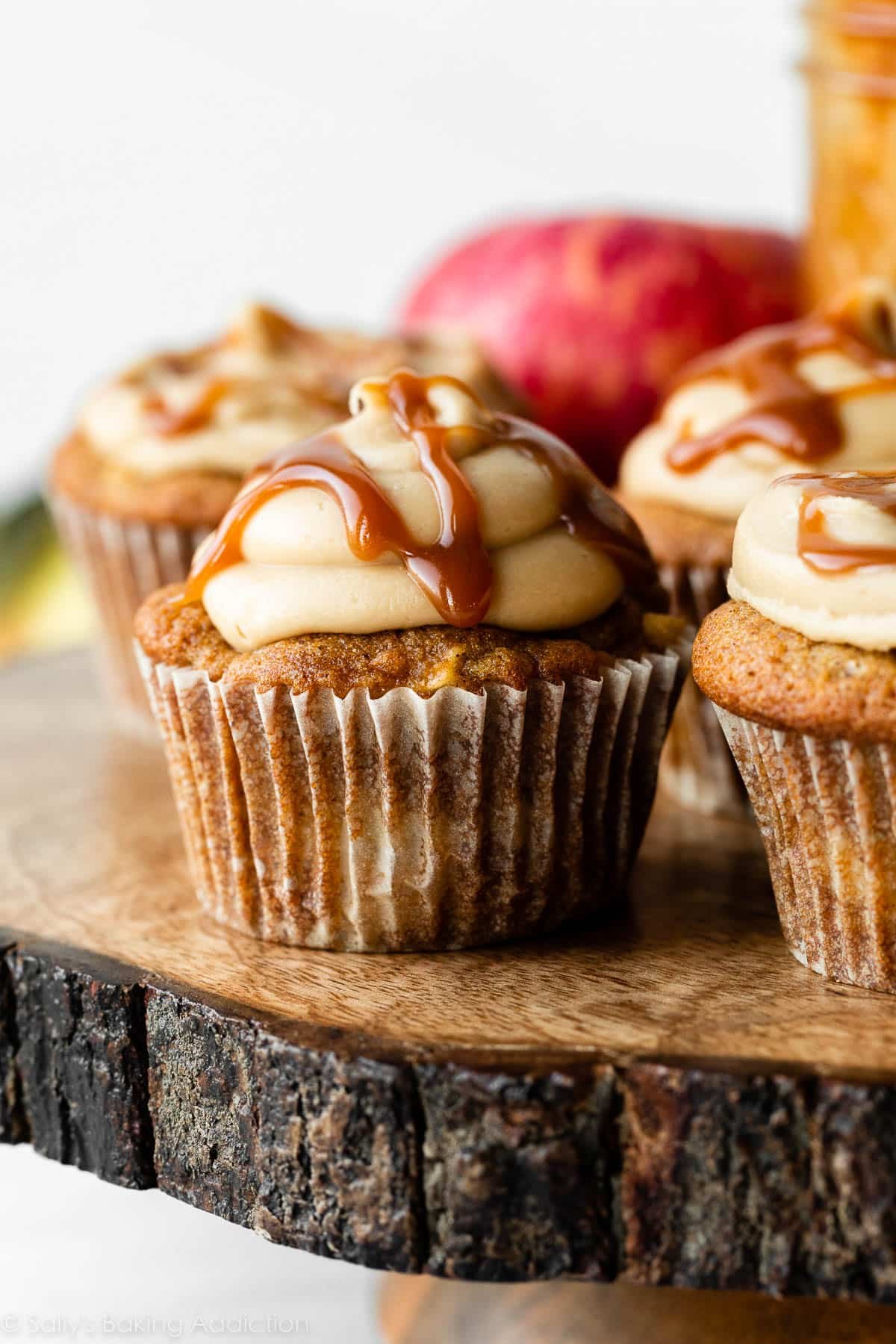 apple cupcakes topped with salted caramel frosting and caramel sauce sitting on wooden cake stand.