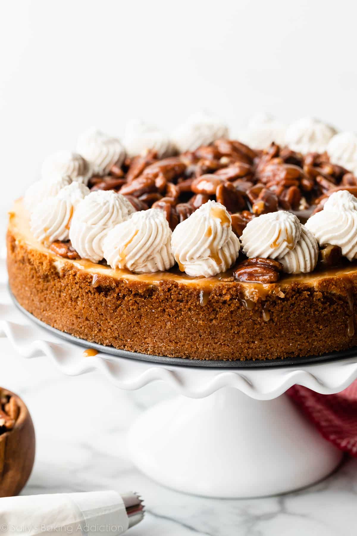 cheesecake on white cake stand topped with pecan pie topping and piped whipped cream around the edges.