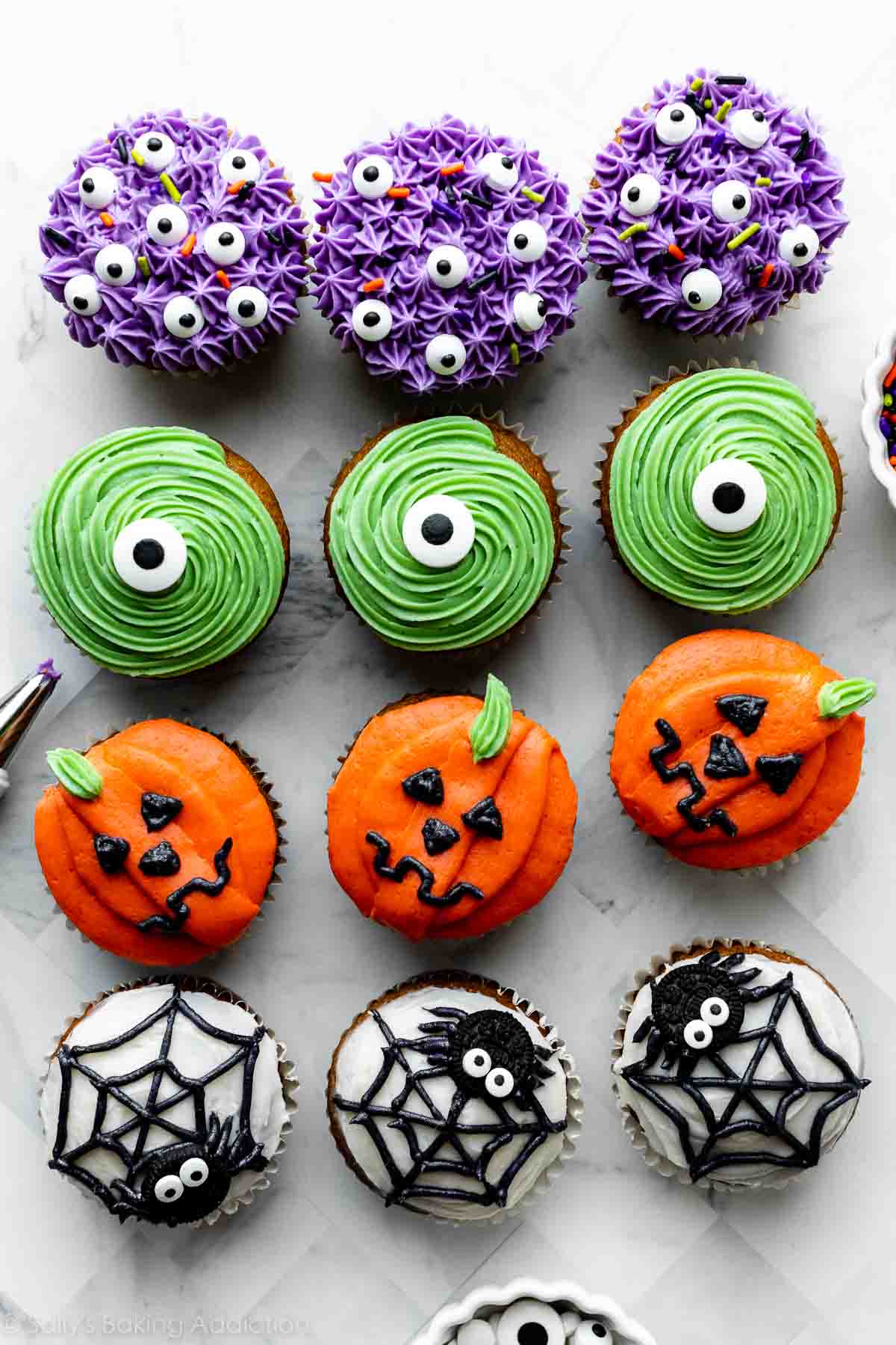 12 decorated Halloween cupcakes including purple monsters, green monsters, spiders and spider webs, and pumpkin Jack-O-Lanterns.