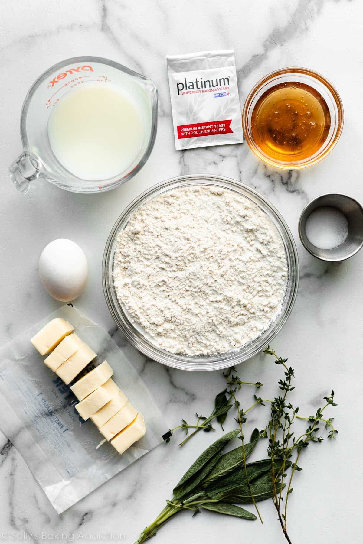 flour, butter, sage, salt, egg, honey, yeast, and milk arranged on marble counter.