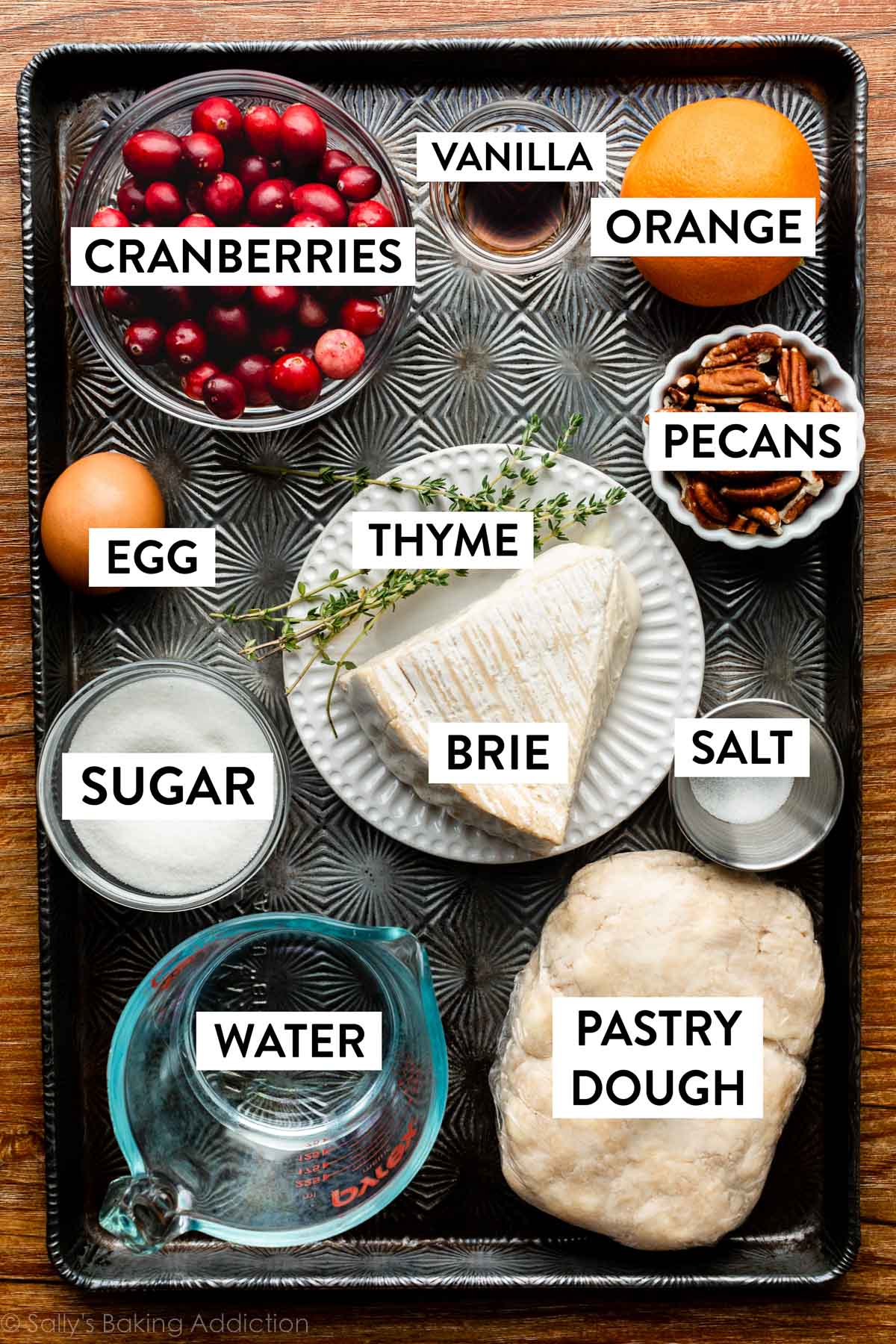 ingredients on dark baking sheet including cranberries, orange, brie, thyme, pastry dough, water, sugar, and pecans.