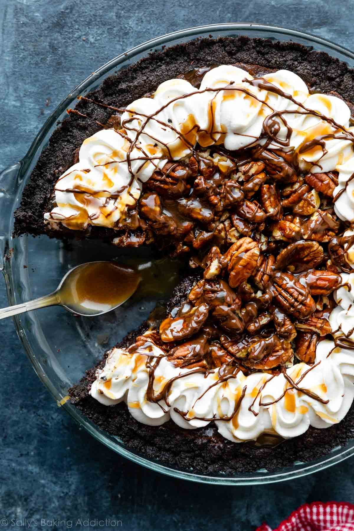 caramel turtle brownie pie topped with melted chocolate, pecans, caramel sauce, and piped whipped cream with slice taken out.