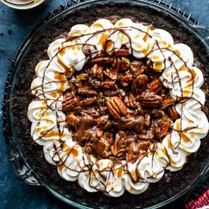 caramel turtle pecan brownie pie on Oreo cookie crust with piped whipped cream around the edges on blue backdrop.