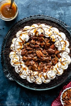 caramel turtle pecan brownie pie on Oreo cookie crust with piped whipped cream around the edges on blue backdrop.