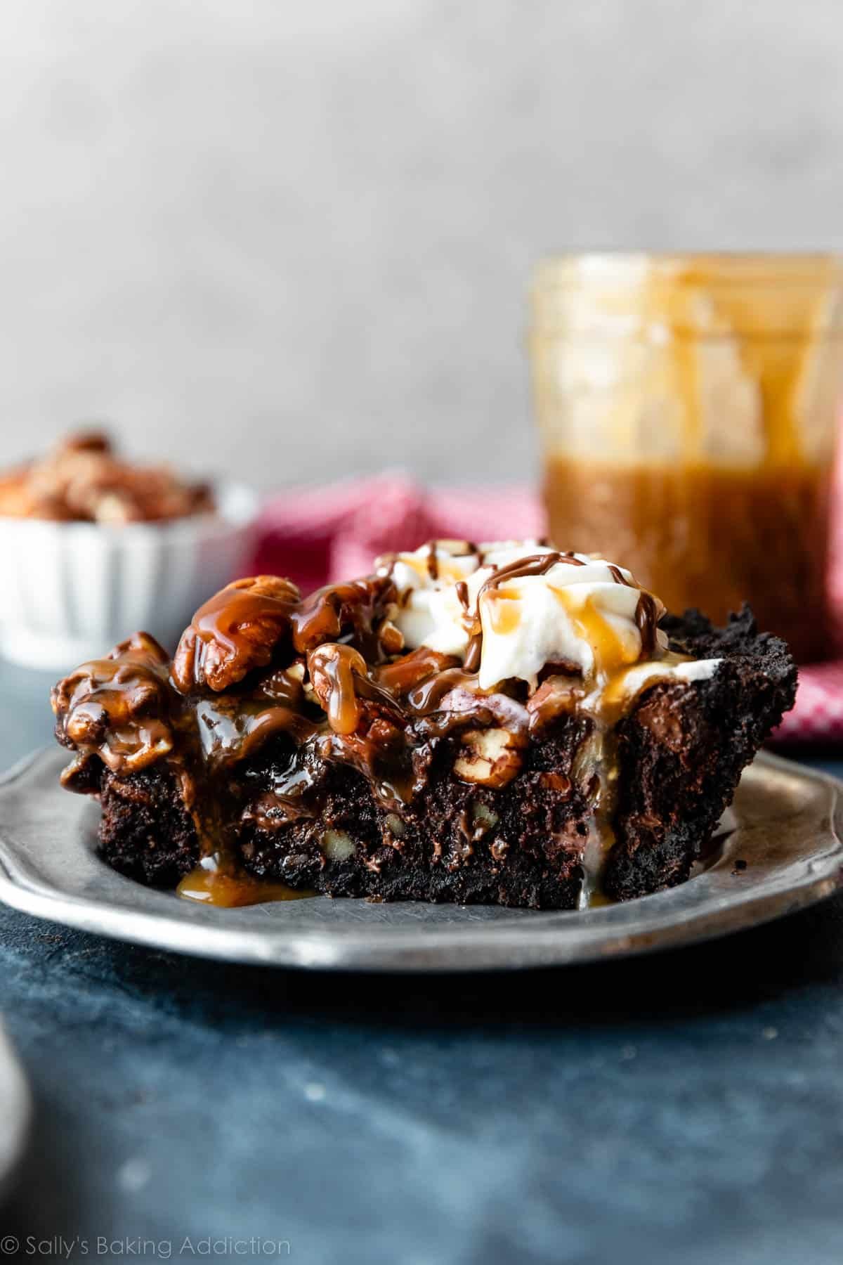 slice of brownie pie with pecans, caramel, and whipped cream on top on a silver plate with jar of caramel in the background.