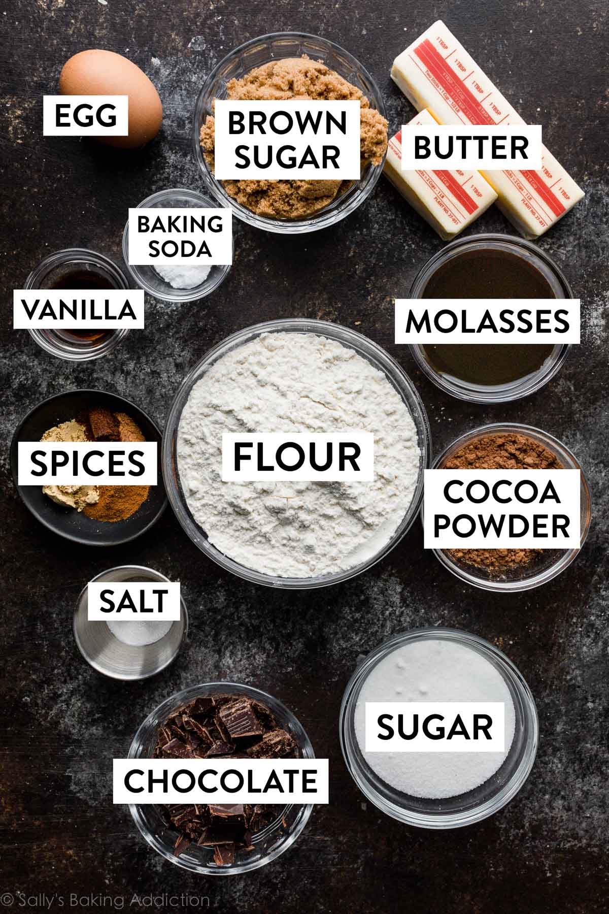 ingredienti sul piano di lavoro grigio nero tra cui ciotole di farina, cacao in polvere, zucchero, cioccolato, sale, spezie, vaniglia, zucchero di canna e melassa.