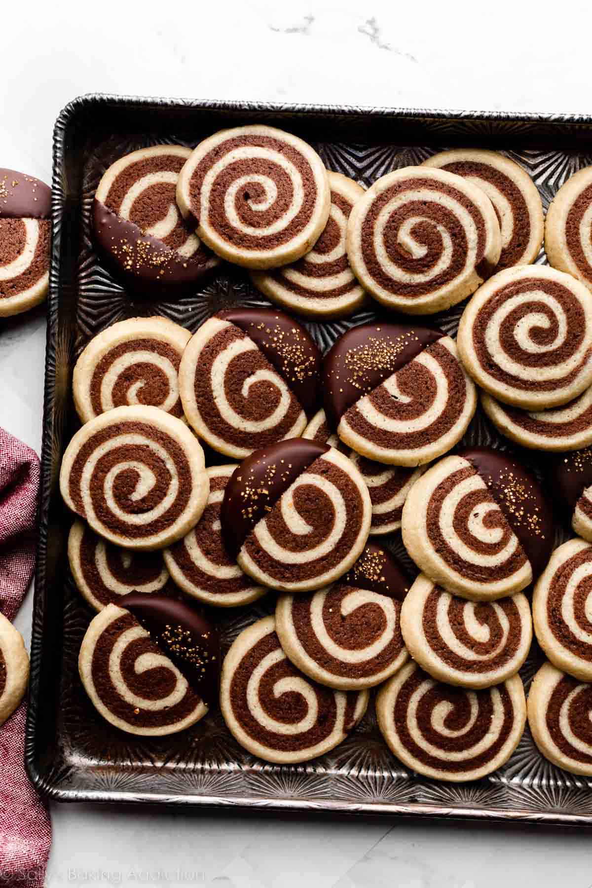pinwheel cookies with some dipped in chocolate and topped with gold sprinkles on dark metal baking sheet.