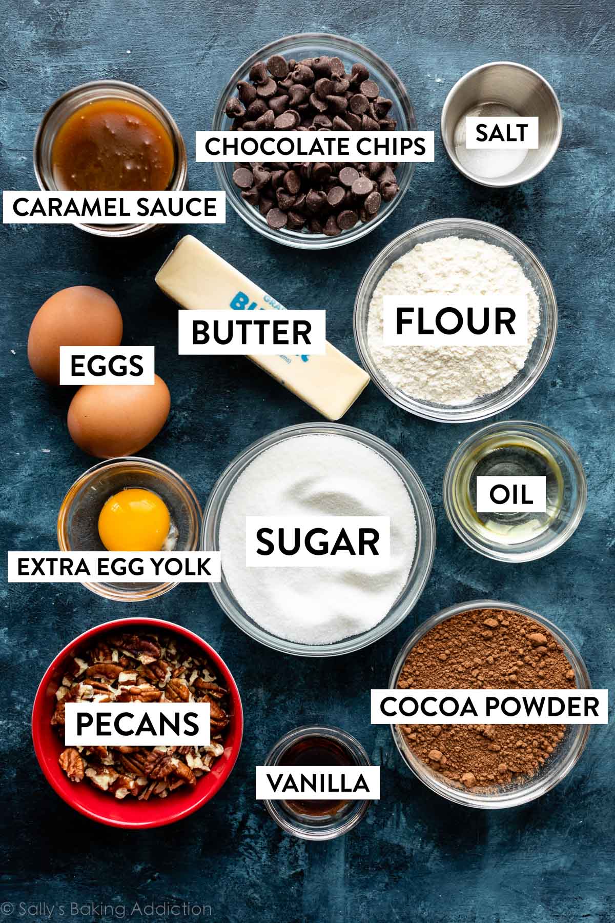 ingredients on blue backdrop including butter, pecans, chocolate chips, flour, eggs, sugar, and caramel sauce.