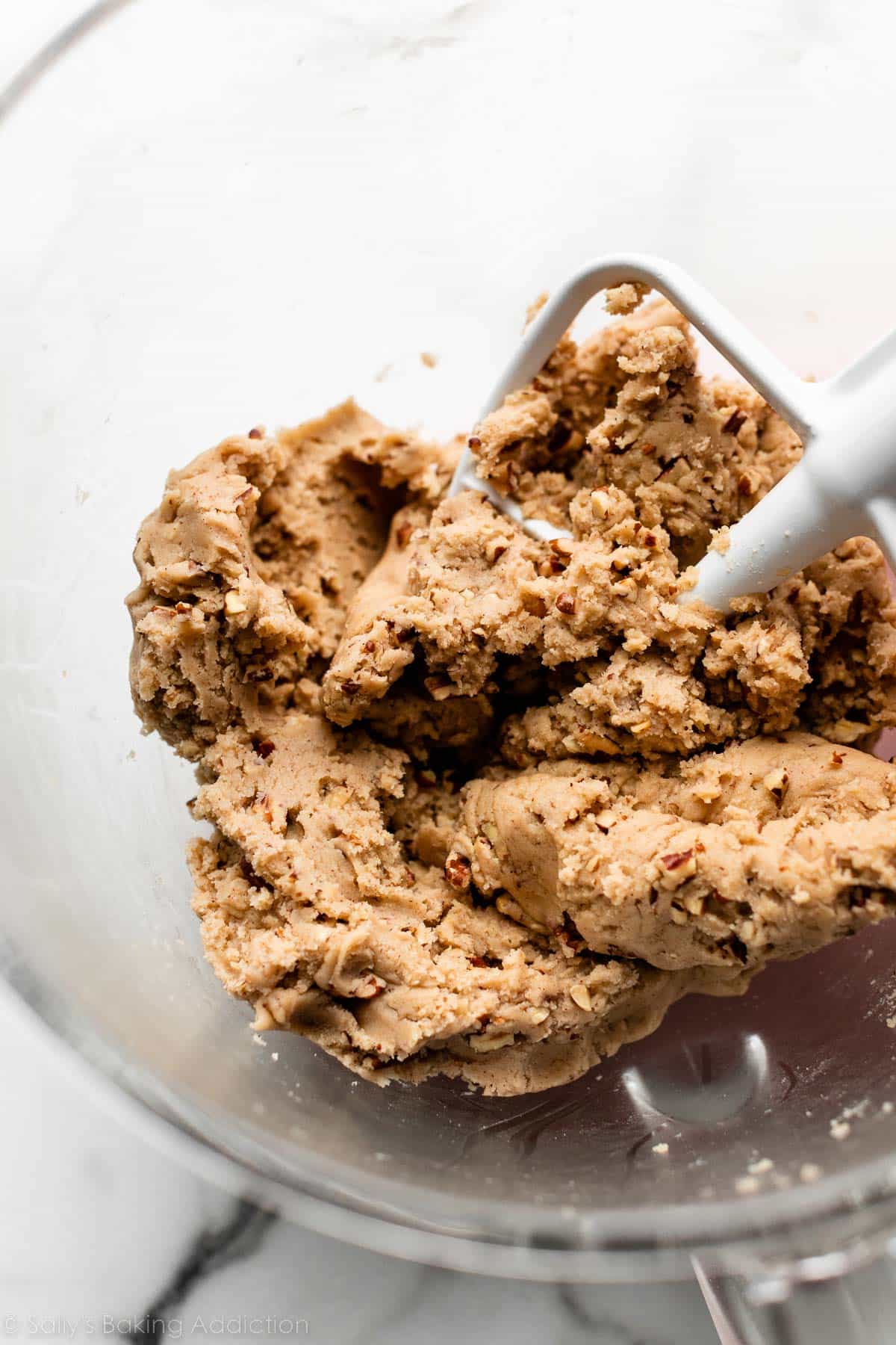 light brown cookie dough filled with finely chopped pecans in glass mixing bowl with paddle attachment.