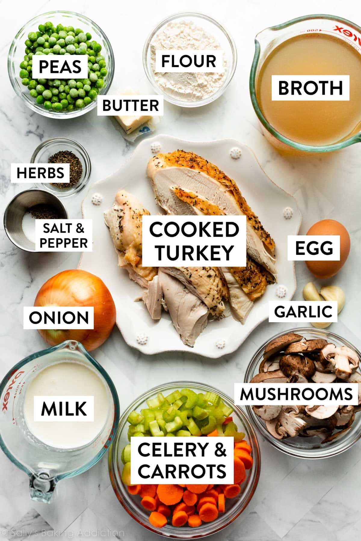 ingredients on marble counter including broth, cooked turkey, peas, mushrooms, milk, garlic cloves, and onion.