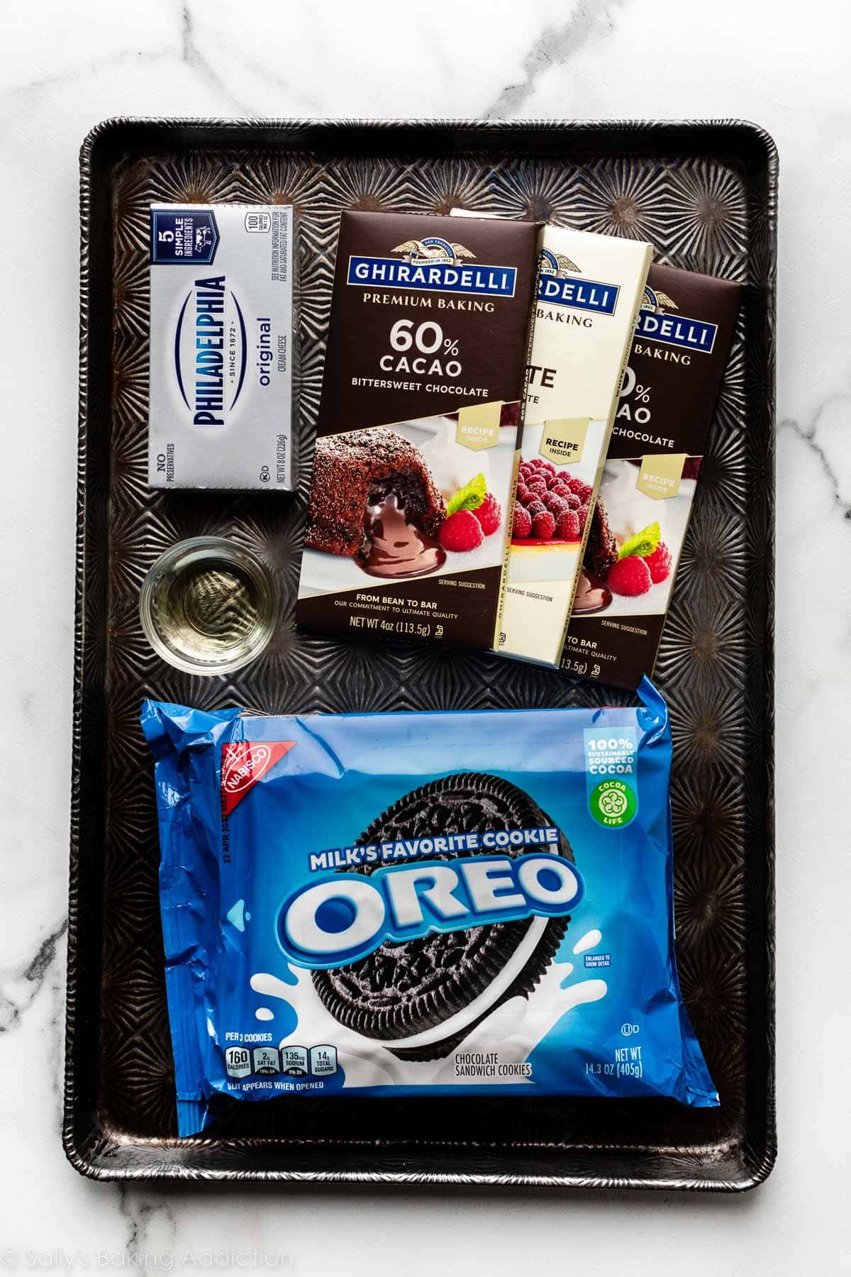 ingredients on baking sheet including chocolate, cream cheese, small bowl of oil, and Oreo cookie package.