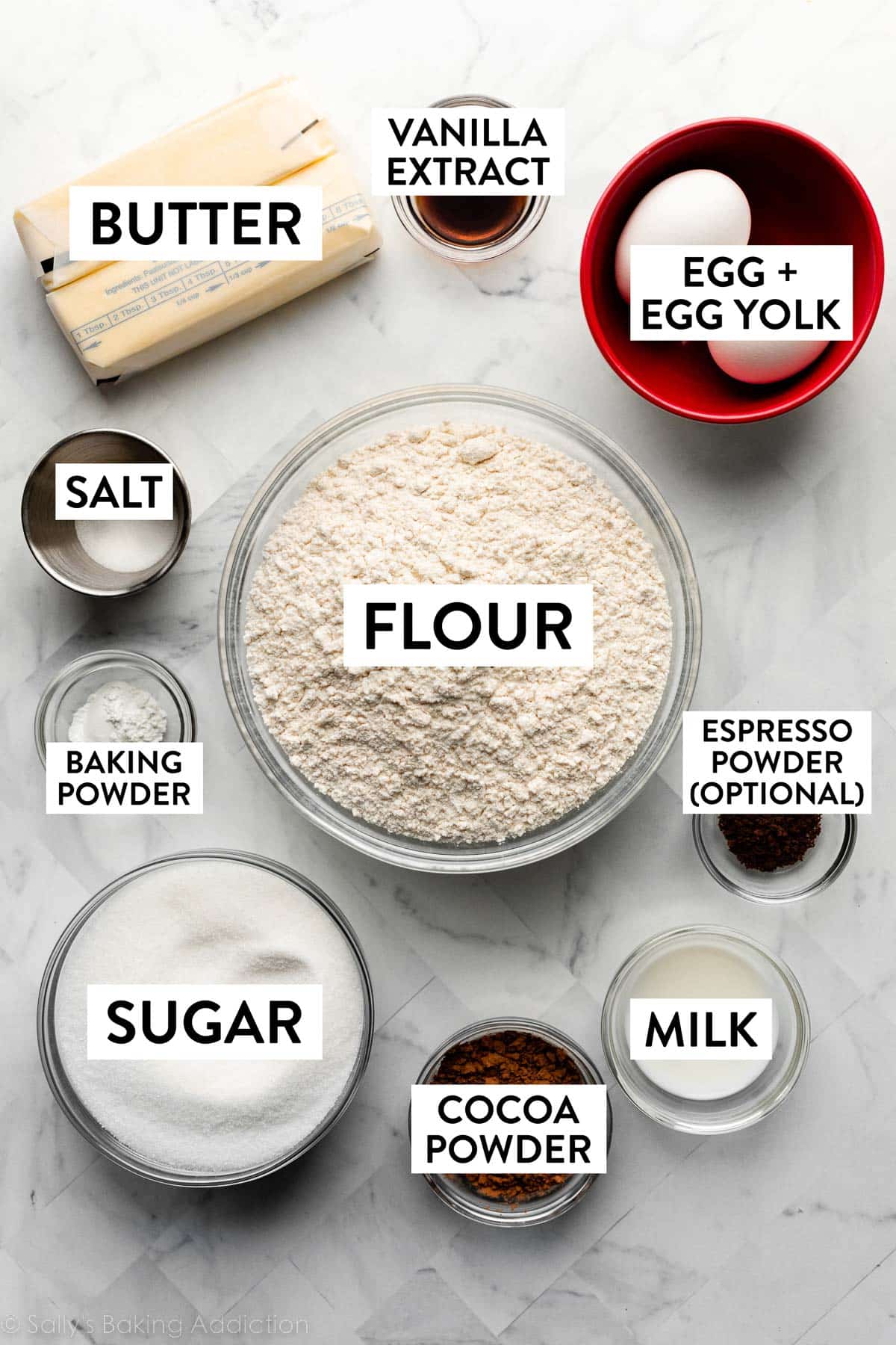 ingredients on marble counter including flour, eggs, sugar, butter, milk, cocoa powder, and vanilla.