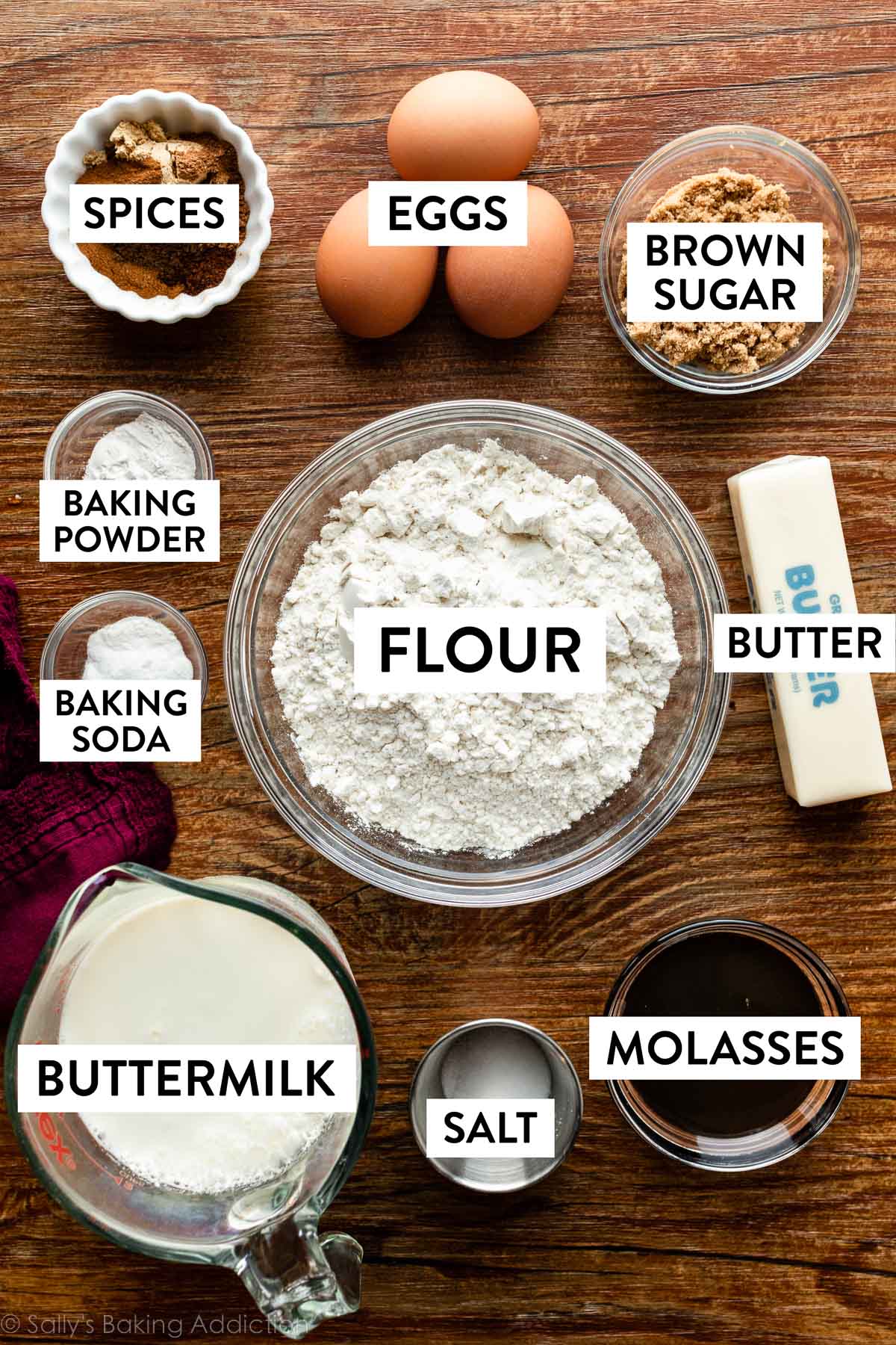 ingredients on wooden backdrop including eggs, flour, molasses, spices, buttermilk, butter, and brown sugar.