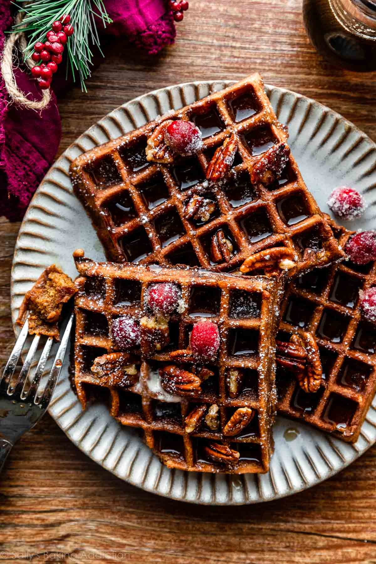 plate of 3 gingerbread waffles with sugared cranberries, pecans, and maple syrup on top.
