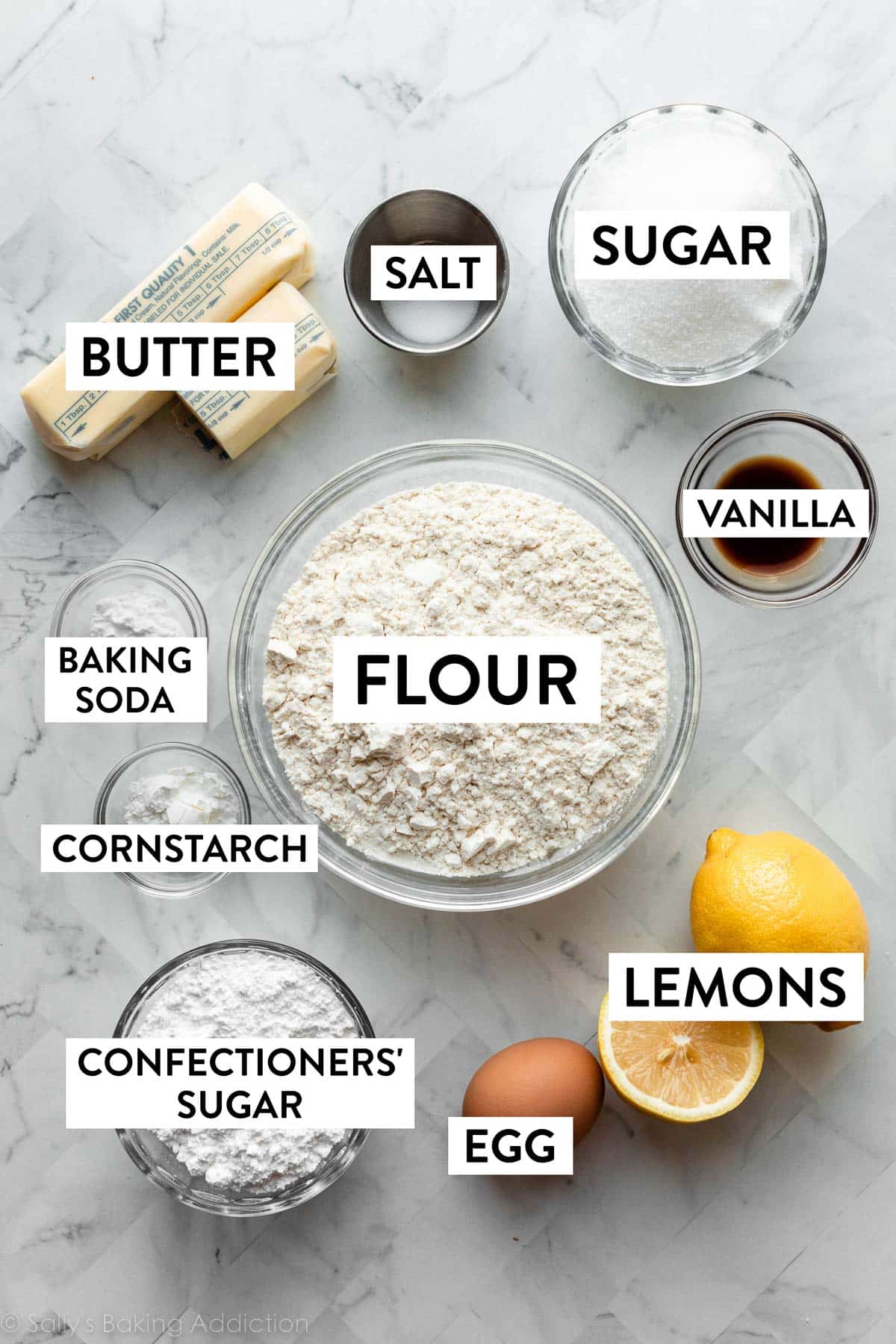 ingredients on marble counter including egg, one and 1/2 lemons, big bowl of flour, sugar, butter, and other ingredients.