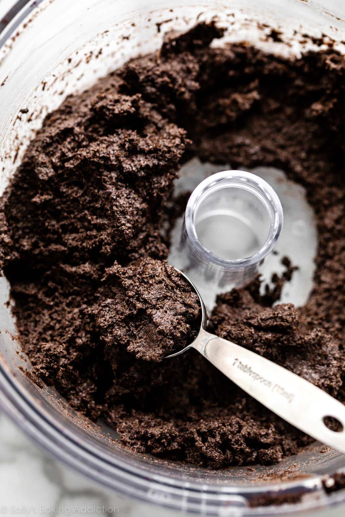 black cookie crumb mixture in food processor with a Tablespoon measuring spoon scooping some out.