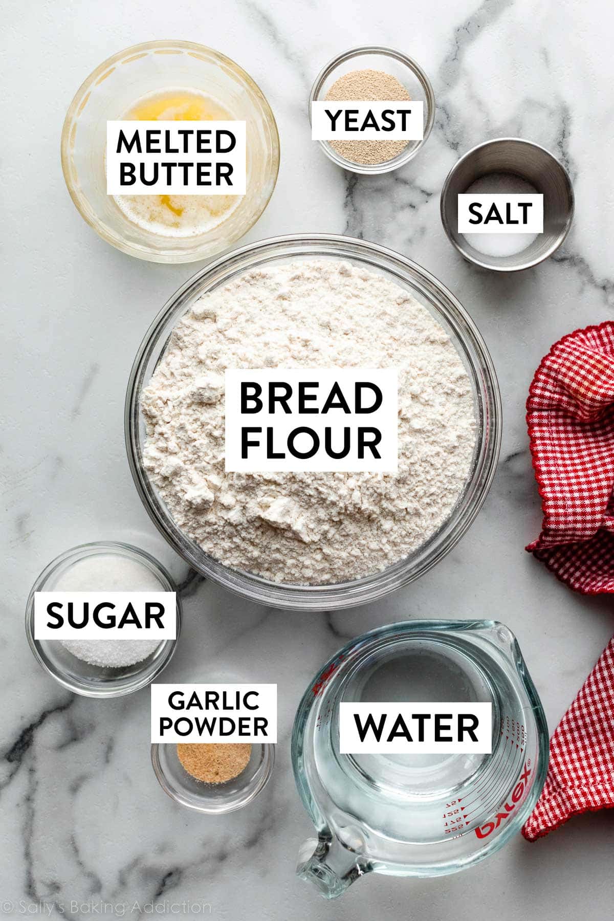 ingredients on marble counter including bread flour, melted butter, yeast, water, garlic powder, salt, and sugar.