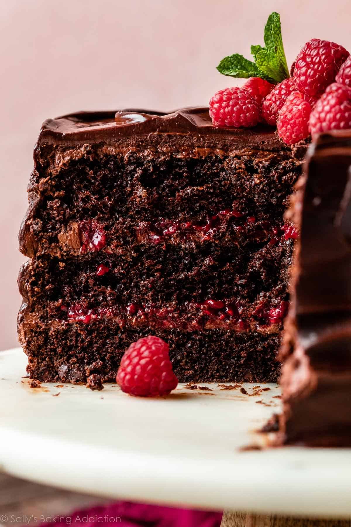 chocolate raspberry cake cut open with raspberries and mint on top sitting on a marble cake stand with pink backdrop behind it.