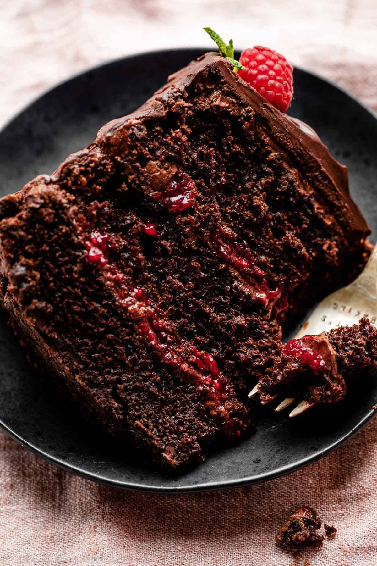 slice of dark chocolate cake with raspberry filling and chocolate ganache on black plate.