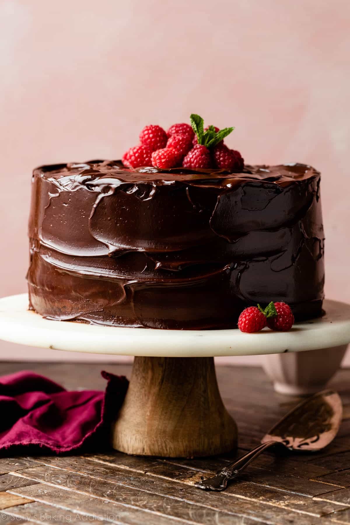 chocolate ganache raspberry cake on marble cake stand with raspberries on top and pink backdrop behind it.