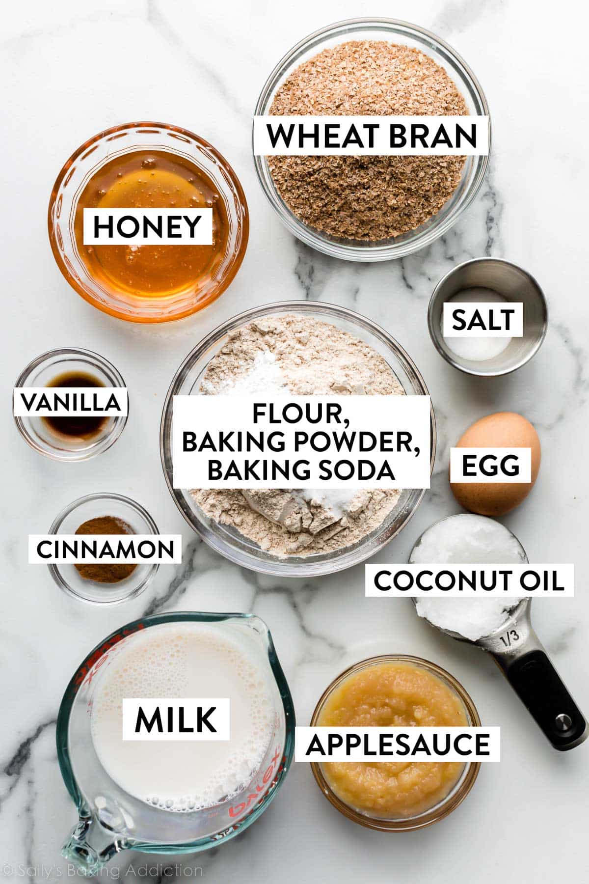 ingredients on marble counter including flour, applesauce, coconut oil, egg, milk, honey, and more.