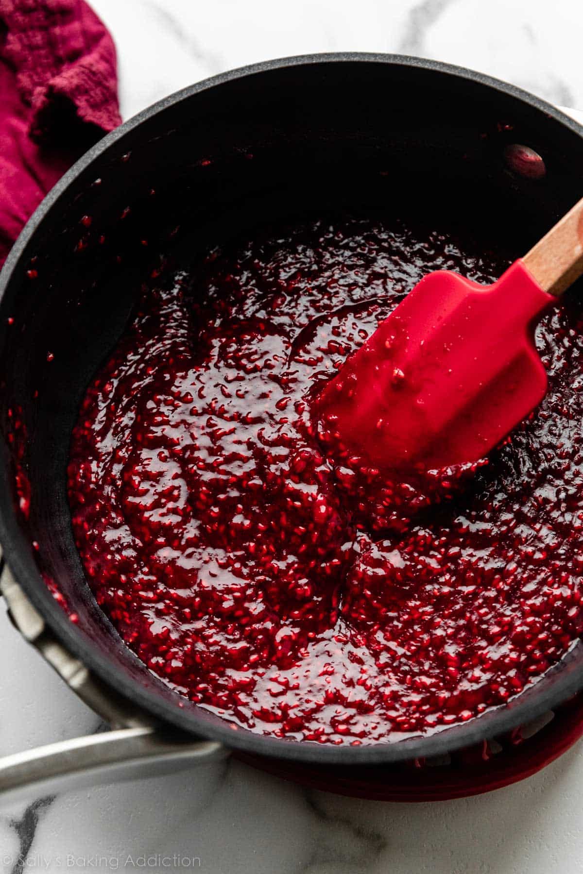 seeded thickened raspberry mixture in black saucepan with red spatula.